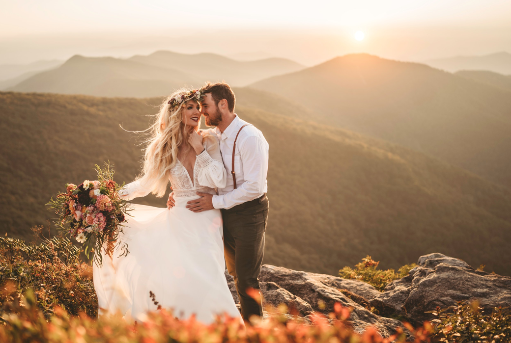 A bride and groom share a joyful moment on a mountaintop during sunset in the Great Smoky Mountains. The bride, wearing a flowing white dress and a floral crown, holds a vibrant bouquet of flowers. The groom, dressed in suspenders and a white shirt, embraces her. The warm, golden light of the setting sun bathes the scene, highlighting the stunning mountain landscape in the background.