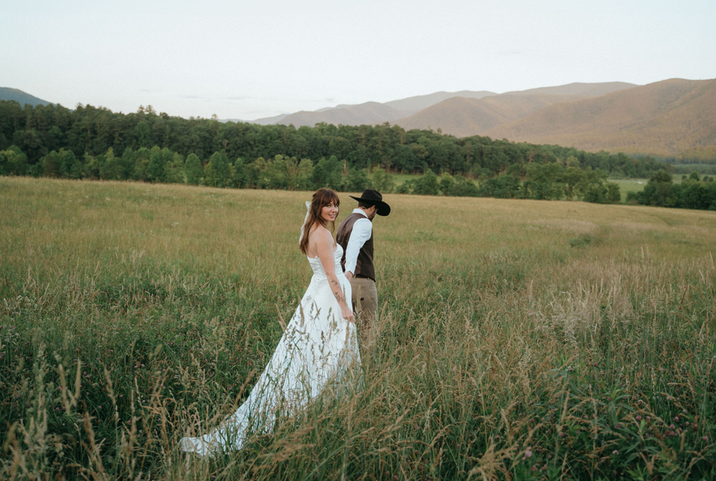 A bride and groom stand in a grassy field with mountains and trees in the background. She's in a white dress, and he's wearing a cowboy hat.