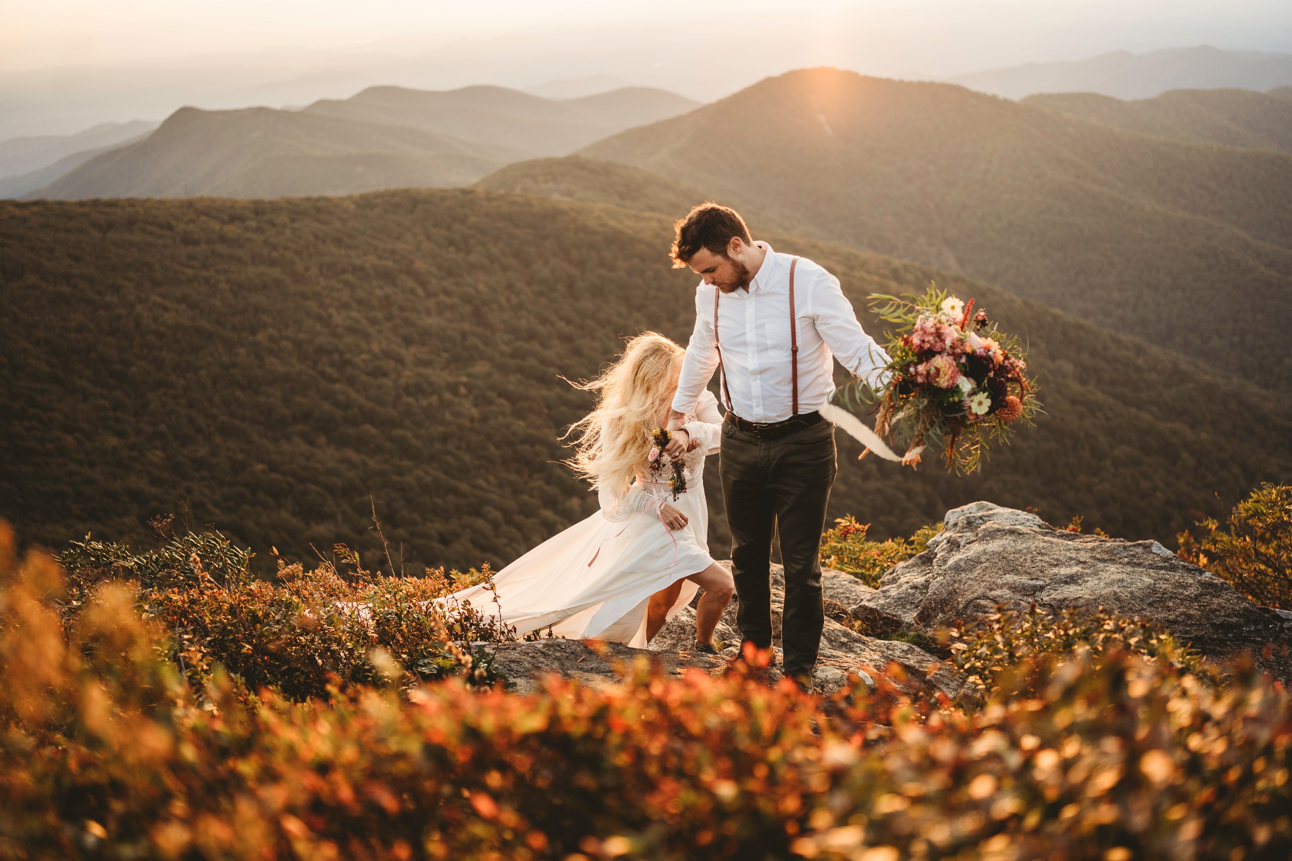 A bride and groom share a joyful moment on a mountaintop during sunset in the Great Smoky Mountains. The bride, wearing a flowing white dress and a floral crown, holds a vibrant bouquet of flowers. The groom, dressed in suspenders and a white shirt, embraces her. The warm, golden light of the setting sun bathes the scene, highlighting the stunning mountain landscape in the background.