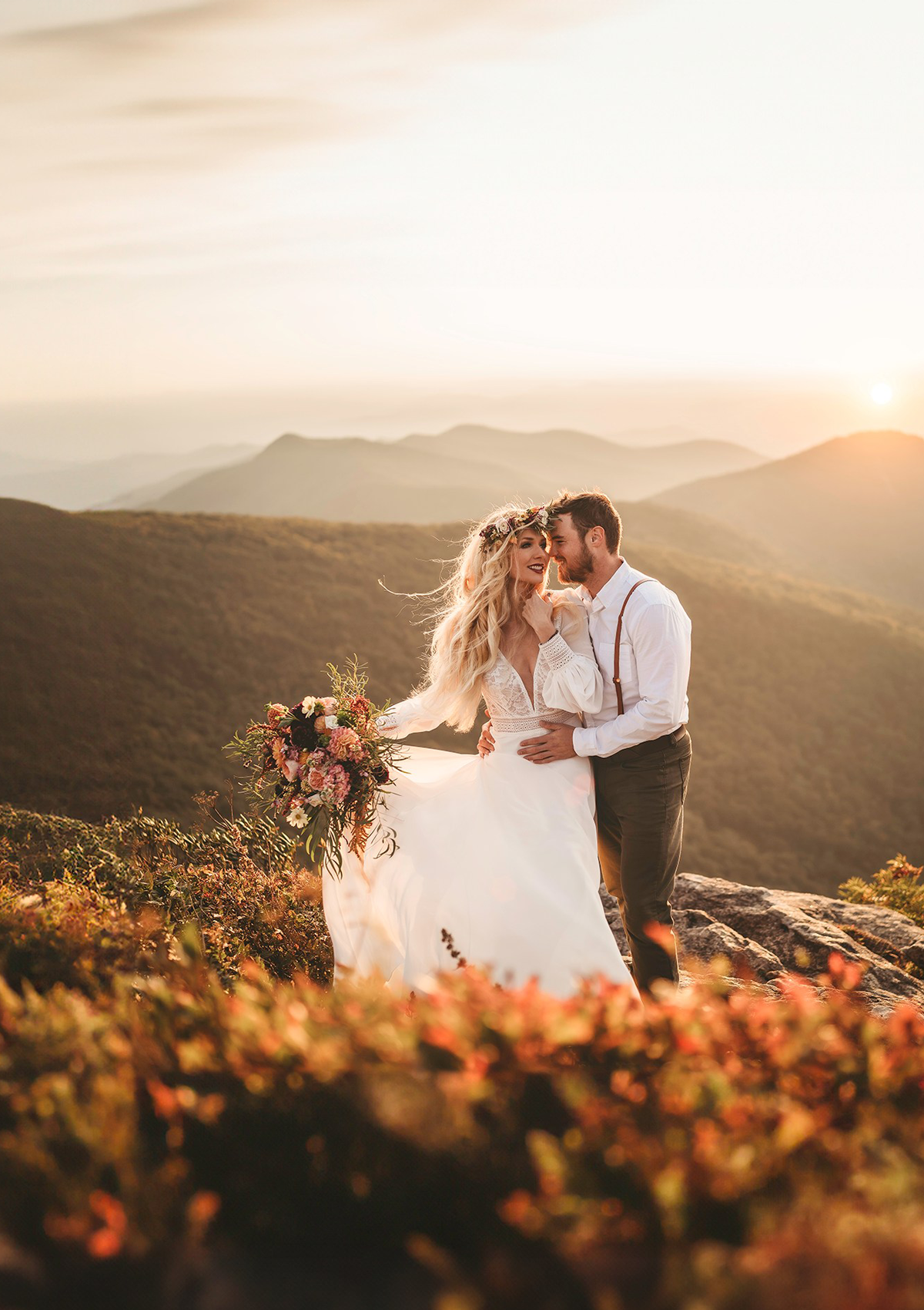 A bride and groom share a joyful moment on a mountaintop during sunset in the Great Smoky Mountains. The bride, wearing a flowing white dress and a floral crown, holds a vibrant bouquet of flowers. The groom, dressed in suspenders and a white shirt, embraces her. The warm, golden light of the setting sun bathes the scene, highlighting the stunning mountain landscape in the background.