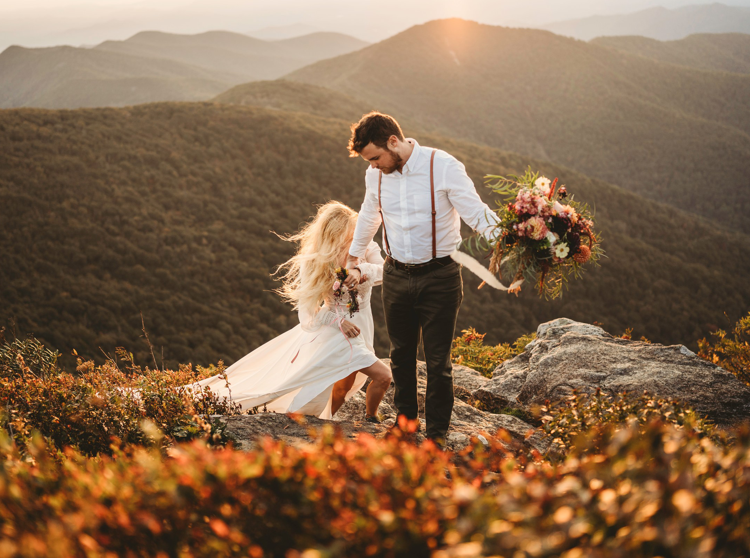 A bride and groom share a joyful moment on a mountaintop during sunset in the Great Smoky Mountains. The bride, wearing a flowing white dress and a floral crown, holds a vibrant bouquet of flowers. The groom, dressed in suspenders and a white shirt, embraces her. The warm, golden light of the setting sun bathes the scene, highlighting the stunning mountain landscape in the background.