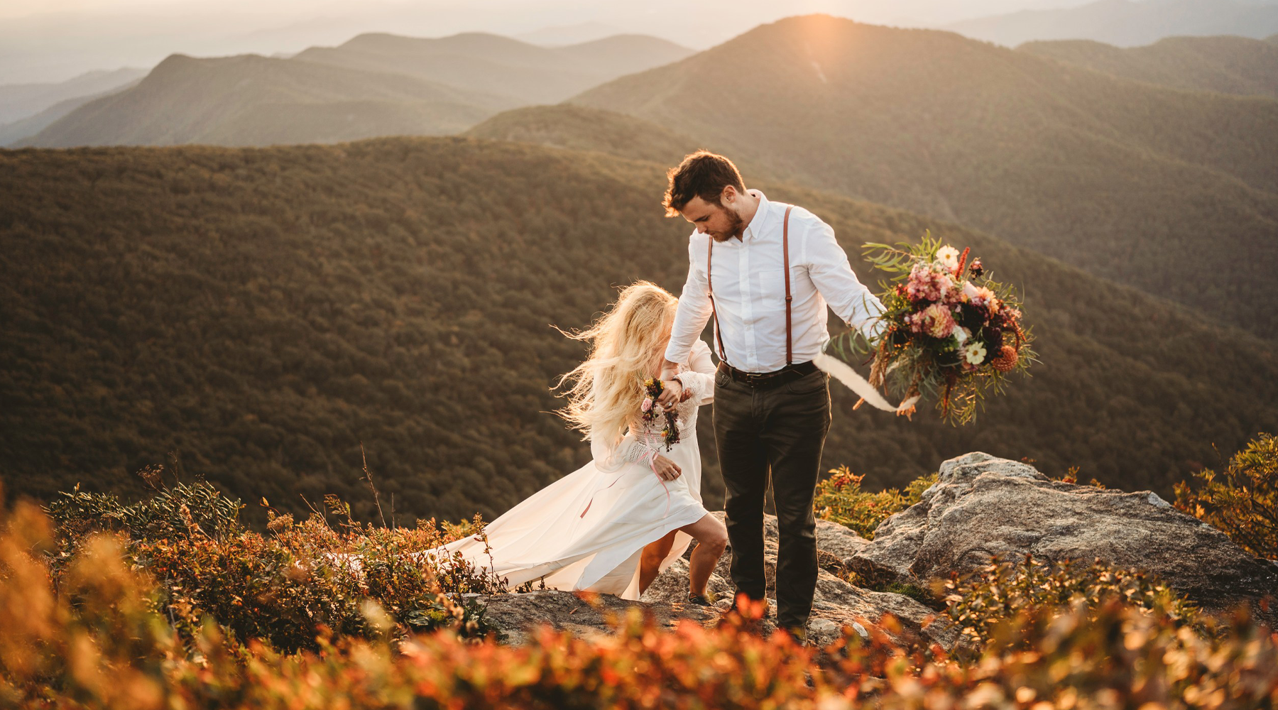 A bride and groom share a joyful moment on a mountaintop during sunset in the Great Smoky Mountains. The bride, wearing a flowing white dress and a floral crown, holds a vibrant bouquet of flowers. The groom, dressed in suspenders and a white shirt, embraces her. The warm, golden light of the setting sun bathes the scene, highlighting the stunning mountain landscape in the background.